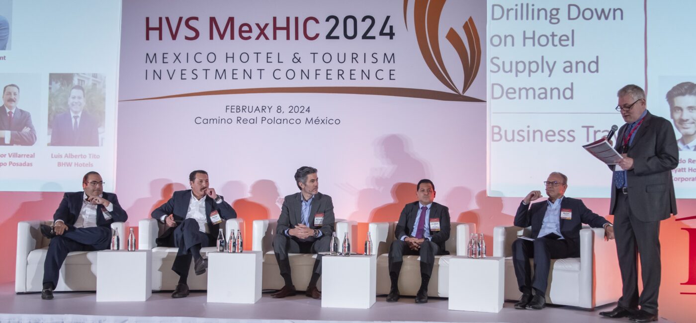 Seis personas con traje sentadas en escenario de panel en HVS MexHIC 2024 conferencia hotelera en México.