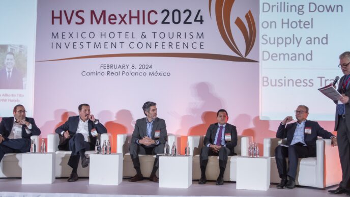 Seis personas con traje sentadas en escenario de panel en HVS MexHIC 2024 conferencia hotelera en México.
