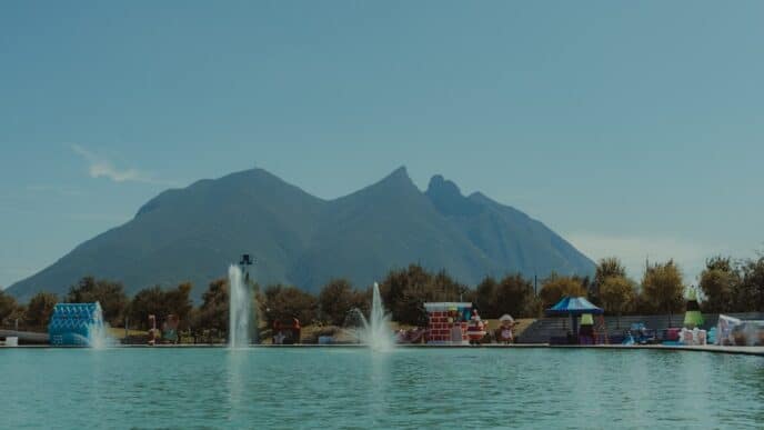 Alberca con fuentes frente a árboles y montañas bajo cielo azul sin nubes.