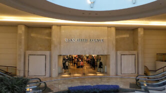 Persona entrando a tienda Saks Fifth Avenue en centro comercial, escaleras eléctricas y plantas al frente.