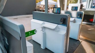 People use kioscos de check-in automático en aeropuerto junto a zona al aire libre con carpas.