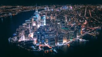 Vista aérea nocturna del skyline de Nueva York junto al agua, calles iluminadas y edificios densos.