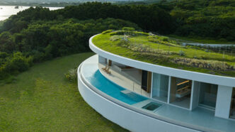 Casa curva con techo verde, alberca y persona en jardín rodeado de vegetación y montañas.