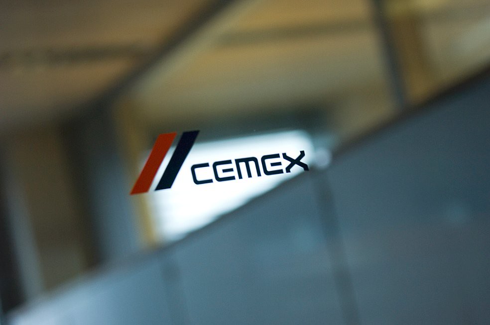 Persona frente a logotipo de Cemex con franjas rojas y azules sobre panel de vidrio en oficina.