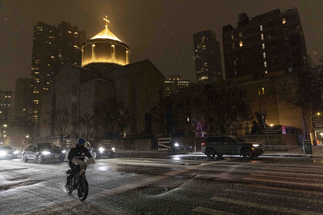 Persona en bicicleta cruza calle nevada de noche, pasan autos, iglesia iluminada y Summit Properties lista peores caseros NYC.