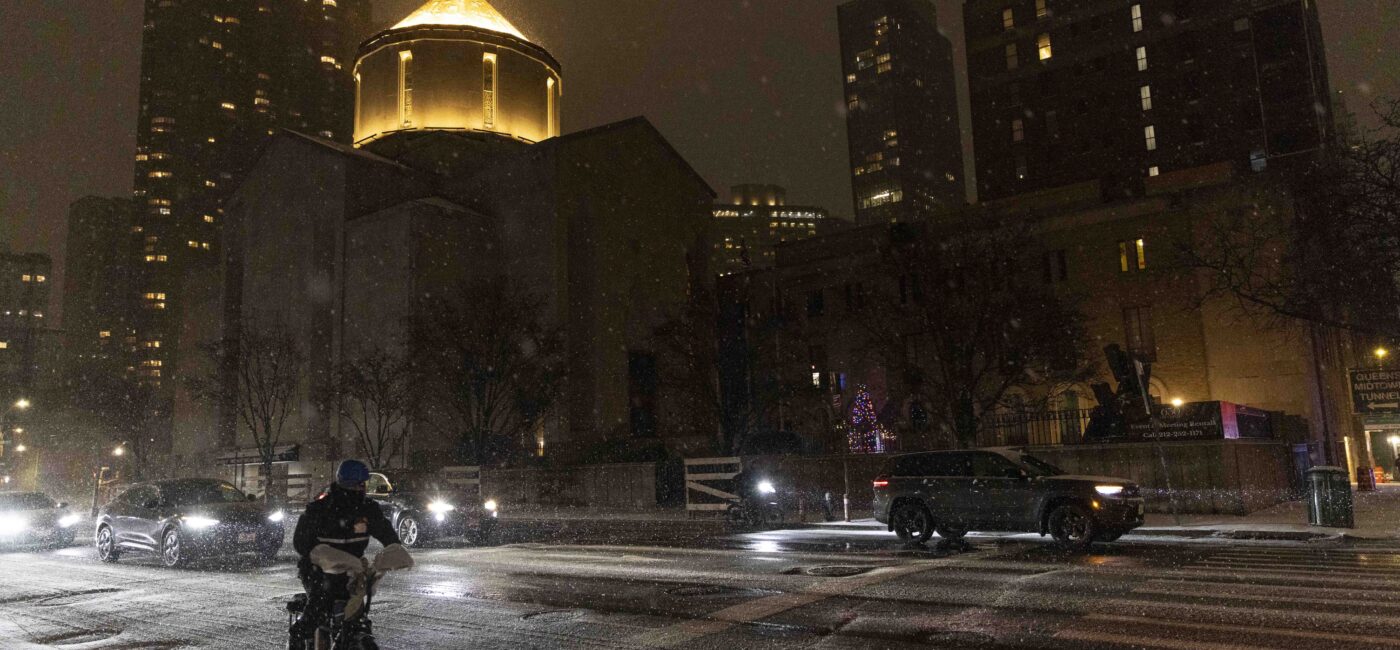 Persona en bicicleta cruza calle nevada de noche, pasan autos, iglesia iluminada y Summit Properties lista peores caseros NYC.