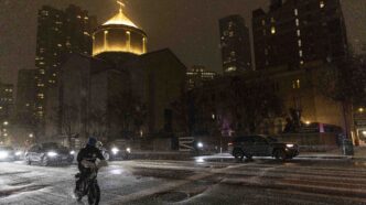 Persona en bicicleta cruza calle nevada de noche, pasan autos, iglesia iluminada y Summit Properties lista peores caseros NYC.
