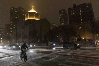 Persona en bicicleta cruza calle nevada de noche, pasan autos, iglesia iluminada y Summit Properties lista peores caseros NYC.