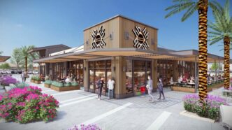 Personas sentadas fuera de restaurante de grandes ventanas entre palmas y flores en plaza comercial.