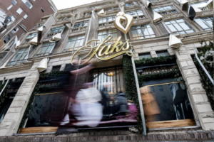 Fachada de Saks con personas difuminadas y anuncio visible de bonos de Saks a 1 centavo en la entrada.