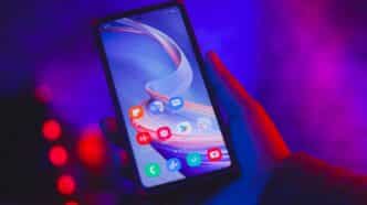 Persona sostiene smartphone mostrando iconos de apps en pantalla, fondo iluminado en tonos morado y azul.