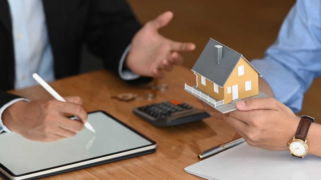 Dos personas conversan en un mercado, una sostiene maqueta de casa y otra escribe en tablet.