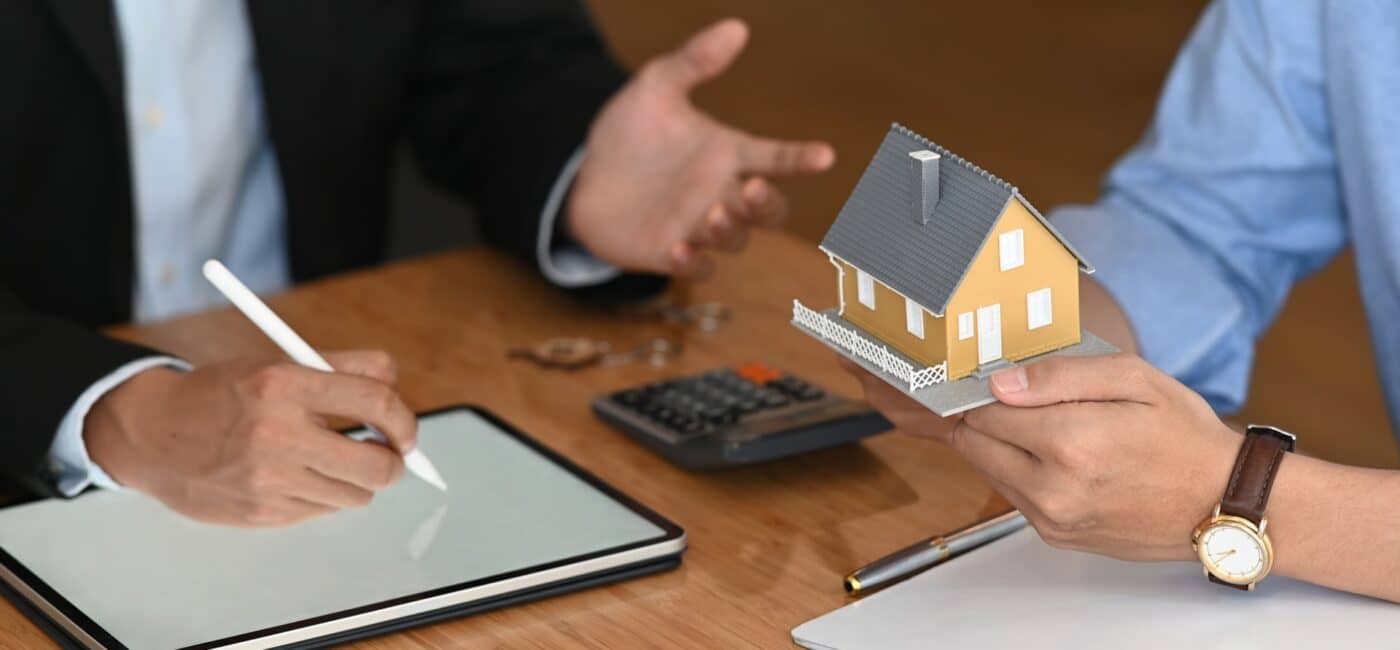 Dos personas conversan en un mercado, una sostiene maqueta de casa y otra escribe en tablet.